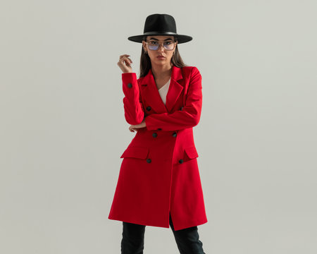 sexy casual girl with hat and glasses holding arms in fashion pose while wearing red coat and posing on grey backgroundの写真素材