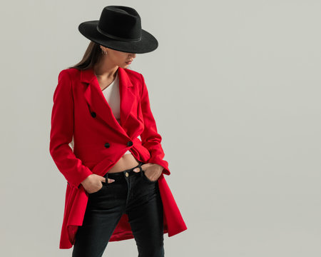 portrait of sexy young woman with long hair and hat holding hands in pockets and looking down side in front of grey backgroundの写真素材