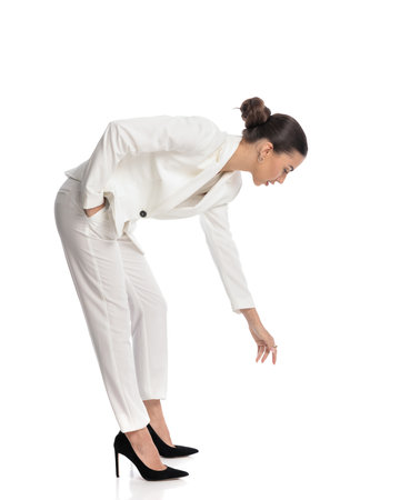 side view of elegant woman in white suit holding hand in pocket, looking down and bending to pick up something from the floor on white backgroundの写真素材