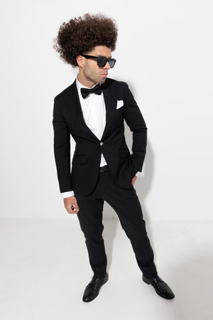 cool fashion businessman in tuxedo holding hands in pockets and looking to side in front of grey backgroundの写真素材