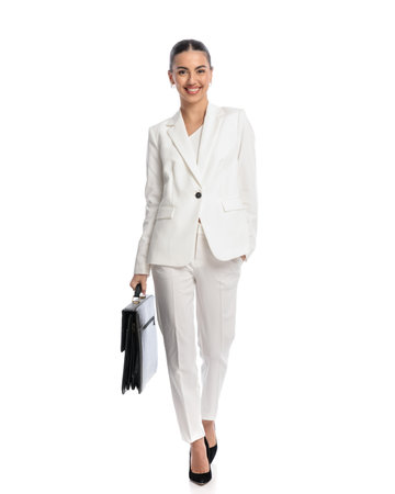 happy confident woman in white suit holding suitcase, smiling and walking with hand in pocket in front of white backgroundの写真素材