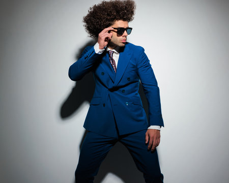 portrait of sexy young businessmn with long curly hair looking to side and fixing sunglasses while confidently posing on grey backgroundの写真素材