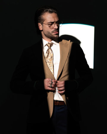 cool young businessman with eyeglasses looking down side and buttoning suit in a mysterious way in front of window shadow on grey backgroundの写真素材