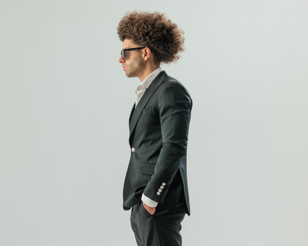 side view of elegant man with afro hair and sunglasses holding hands in pockets and looking to side on grey backgroundの写真素材