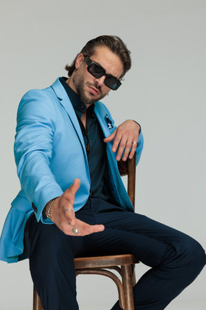 elegant fashion businessman with sunglasses sitting and making hand gesture, pointing hand forward on grey backgroundの写真素材