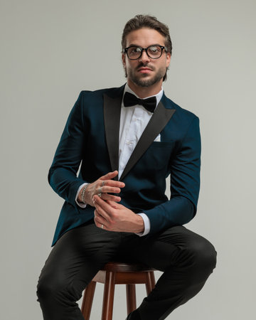 portrait of handsome elegant man with glasses looking forward and adjusting rings while sitting in front of grey backgroundの写真素材