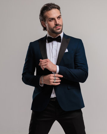 confident young businessman in tuxedo looking to side and adjusting sleeves while posing in front of grey backgroundの写真素材