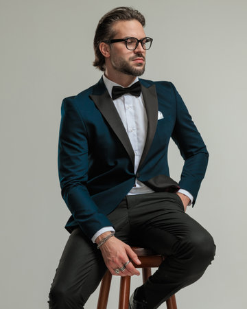 attractive high class businessman with glasses looking to side and sitting with hand in pocket in front of grey background in studioの写真素材