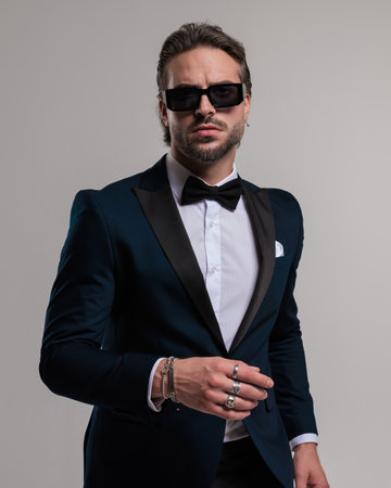 cool fashion man with sunglasses wearing tuxedo and looking forward while posing in a confident way on grey backgroundの写真素材
