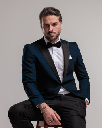 sexy bearded groom in elegant tuxedo looking forward and sitting with hand in pocket in front of grey backgroundの写真素材