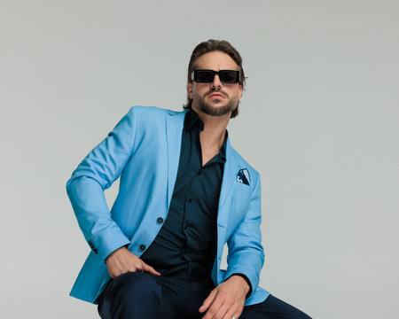 cool fashion man in blue suit with sunglasses sitting and posing with arms on thighs in front of grey backgroundの写真素材