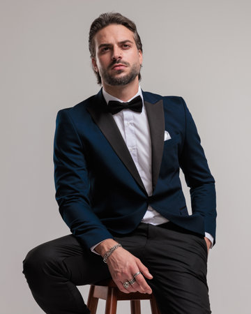 confident high class best man looking up and holding hand in pocket while sitting in front of grey backgroundの写真素材