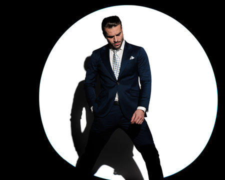 elegant businessman in navy blue suit looking down and posing with hand in pocket in front of white background with spotlight onの写真素材