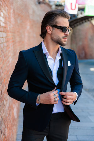 handsome elegant man with glasses looking to side and buttoning suit while posing outdoor in an old city from Romaniaの写真素材
