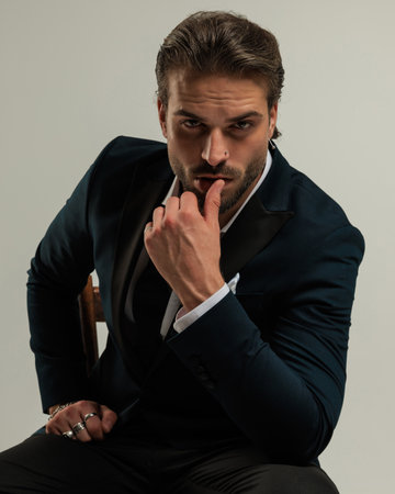 seductive high class man in tuxedo looking forward and sensually touching lips while sitting in front of grey backgroundの写真素材