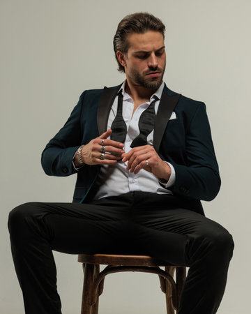cool fashion businessman in tux with undone bowtie sitting and looking down while touching fingers in front of grey backgroundの写真素材