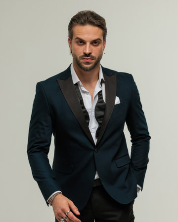 attractive elegant man with beard wearing tuxedo with open collar shirt holding hand in pocket and walking in front of grey backgroundの写真素材