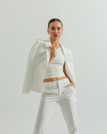 portrait of sexy woman in white suit adjusting jacket, looking forward and posing with hand in pocket in front of grey backgroundの写真素材