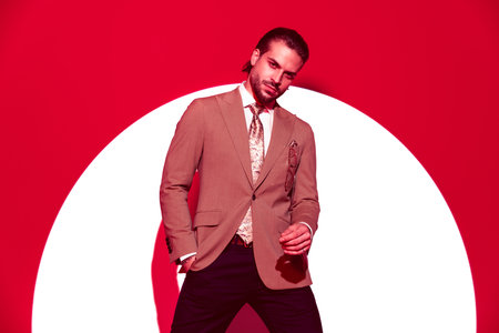 portrait of cool young man in suit holding hand in pocket and looking forward while posing in a confident way in front of spotlight with red lightsの写真素材