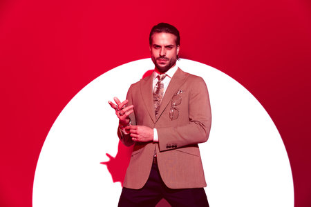 elegant high class businessman looking forward and adjusting bracelets while posing in front of red spotlight backgroundの写真素材