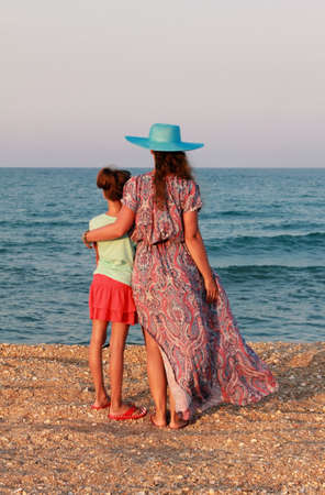 Young mother in blue hat with daughter sister on the sea coastline looking far ahead. windy sunset. Golden hour.の写真素材