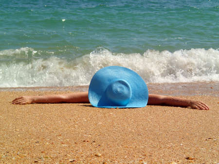 Young woman girl in blue hat laying on the sea beach hiding her face. windy sand sunny weather.の写真素材