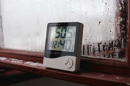 Electronic clock, calendar, thermometer, and hygrometer, against the background of condensation on glass, high humidity. Digital hygrometer in the laboratory shows the temperature of the pharyngeiteの写真素材