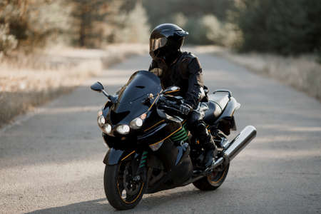 Motorcycle driver in a leather suit and sitting on a motorcycle alone on an asphalt highway against a forest backgroundの写真素材