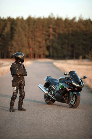 Motorcyclist in leather outfit and a helmet stands on the road and looks at a sports motorcycleの写真素材