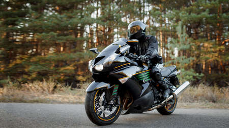 Motorcycle driver in a leather suit and sitting on a motorcycle alone on an asphalt highway against a forest backgroundの写真素材