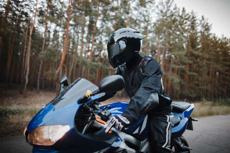 Motorcyclist in leather protective suit and black helmet sits on sports motorcycle. Biker in black rides on the road against the background of the forestの写真素材