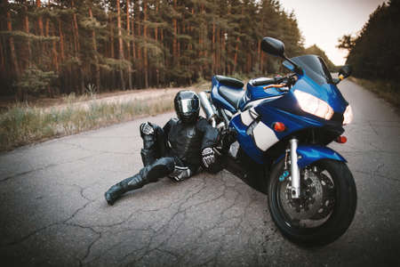 Biker in a protective suit and a black helmet sits next to his motorcycle on the road. Motorcyclist resting near a motorcycleの写真素材