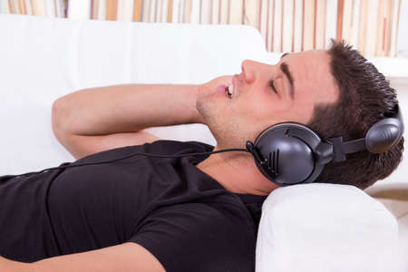 young casual man lying on couch listening to music at home in living room on headphonesの写真素材