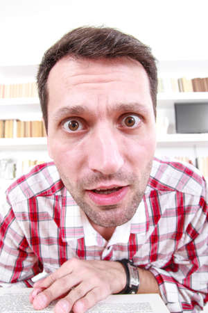 close up of nutty confused and shocked professor or student with book in his hands, living room atmosphere with library backgroundの写真素材