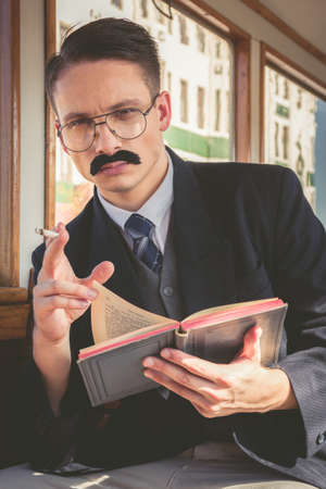 photo of man with glasses and whiskers  in suit sitting in an old wooden wagon train and smoking cigar while reading book , retro vintage fashion styleの写真素材