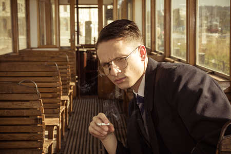 man with glasses in a suit sitting in an old wooden wagon train and smoking a cigar, retro vintage fashion photoの写真素材