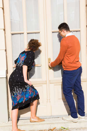 man with glasses in the orange sweater with woman  trying to open the door while looking through the window and curtain , couple buys a houseの写真素材