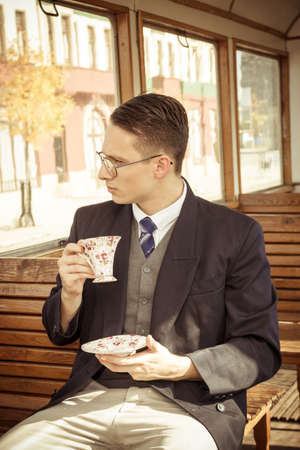 photo of man with mustache and glasses on train wooden wagon drinking coffee or tea from cup and looking through window , retro vintage fashion photoの写真素材