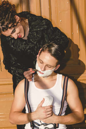 man with shaving foam on his face and towel around his neck siting on chair and  posing  while pretty smiling women shave him , retro vintage fashion compositionの写真素材
