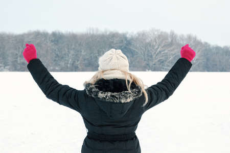 winter woman in snow showing her back and facing forest with fist hands raised high above, copy space outside on cold winter day. Portrait Caucasian happy joyful female model with pink gloves and hat in first snowの写真素材