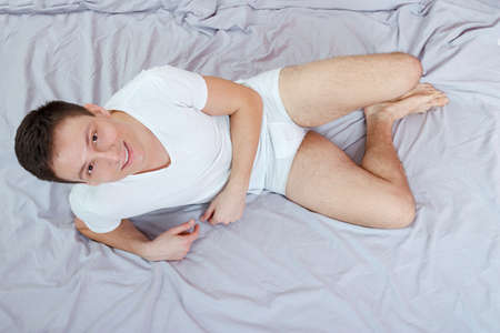 Handsome smiling caucasian young man in white t-shirt and underwear lying or sitting on a bed. Isolated over gray sheet. Domestic atmosphere photo. Sexy attractive male lover in bedroom. Top viewの写真素材