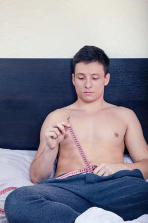 Young man holding tape measure length or size while sitting on bed in bedroom in pajamas. の写真素材