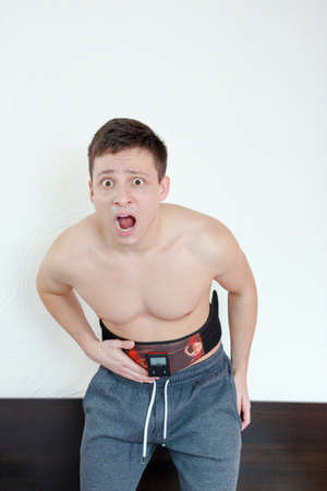 Shocked and stressed handsome young man with an electrical device around his waist works in simple and easy way crunches and removes fat from stomach. Success without practicing sit-ups,six packs abs.の写真素材