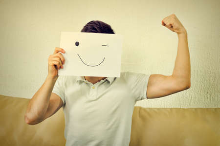 Boy winks and raises fist. Young male advertises successful model body. Man cover his face with happy smile drawn on paper.Fanny crazy  photo, man showing poor muscular biceps arm. Vintage retro styleの写真素材