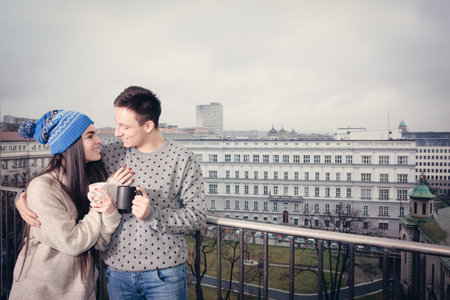 Couple happy in love hugging near railing with cups of tea or coffee. Man and woman drinking hot beverage outdoor on balcony in winter or autumn in cardigan or wool  sweater. Fashion lifestyle photo.の写真素材