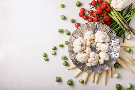 Fresh greens ready to cook: Brussels cabbage, asparagus, baby corn, cauliflower and bamia with metal steamer over a white background. Top viewの写真素材