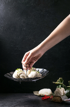 Female hand holding metal steamer with chinese style dumplings with chives served with traditional Japanese onigiri, soy sauce, sweet chili sauce and decorated with banana leaves, chili peppers and chives. Top Viewの写真素材