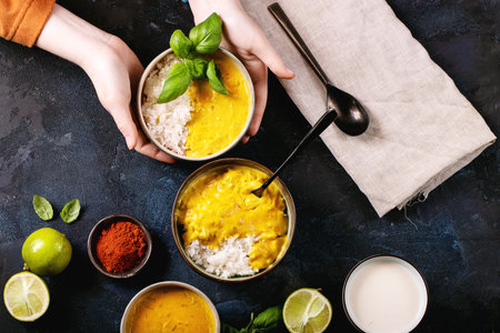 Female holding ceramic bowl of yellow curry served with basil and limes over dark background. Top vew, flat lay. Copy spaceの写真素材