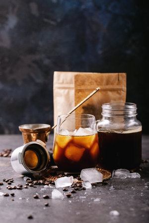 Glass of ice coffee with ice cubes served with cream, copper cezve and coffee beans over dark...