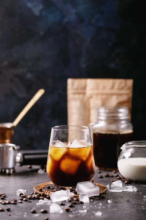 Glass of ice coffee with ice cubes served with cream, copper cezve and coffee beans over dark texture backgroundの写真素材